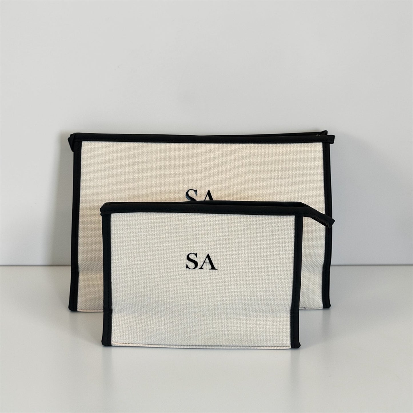 Two beige pouches with black edges and 'SA' monogram on a white background